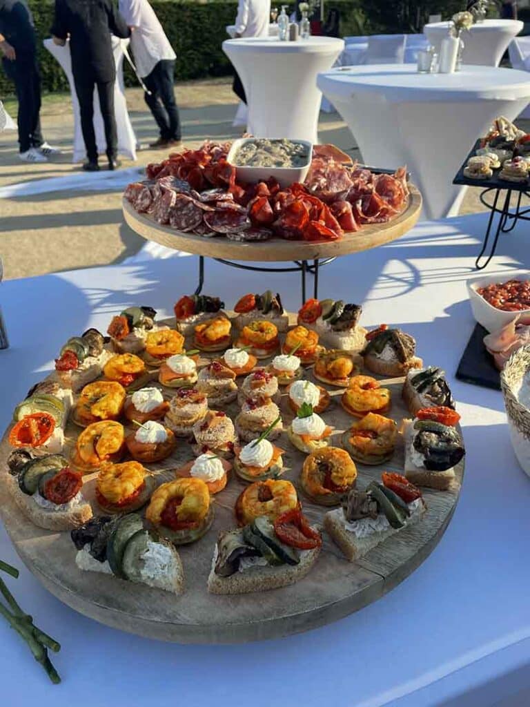 Buffet Traiteur Extérieur : Amuse-bouches et Charcuterie Buffet de réception avec amuse-bouches variés, crevettes et charcuterie sur plateaux en bois en extérieur.