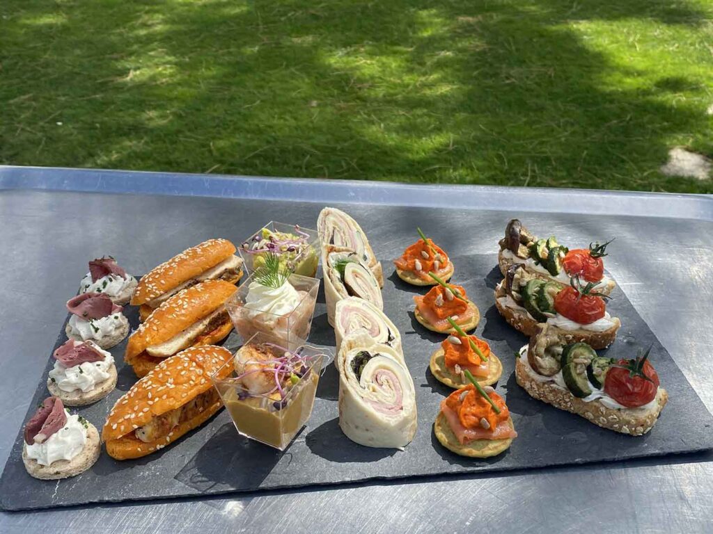Buffet apéritif traiteur : assortiment de canapés gourmands Assortiment apéritif varié avec wraps, mini burgers et bruschettas colorées servis sur une ardoise en extérieur.