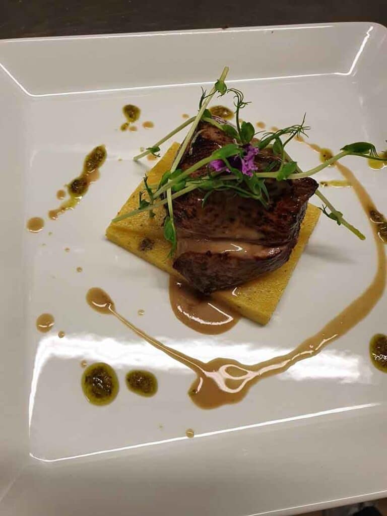 Filet de bœuf grillé sur polenta avec micro-pousses et sauce raffinée dans une assiette carrée.