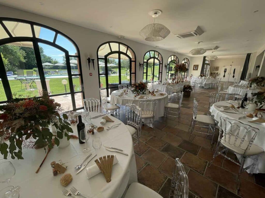 Salle de Réception de Mariage Élégante avec Vue Jardin Réception de mariage élégante : tables dressées avec fleurs et chaises transparentes devant de grandes fenêtres arquées.